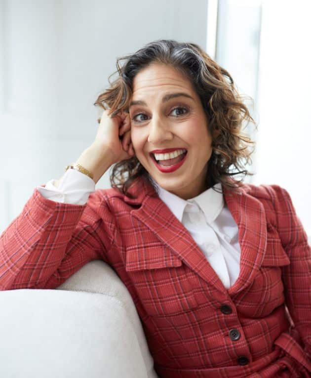 Annahid sitting on a white couch in a red blazer, looking at the camera and smiling.